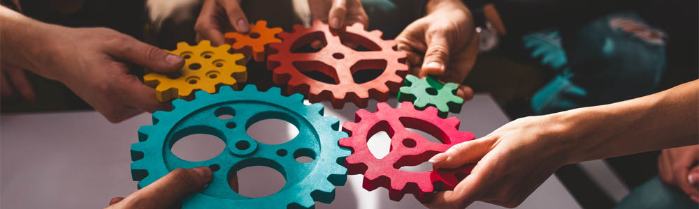 Hands of various people reaching into the middle, all holding different coloured gears and piecing them together like a puzzle
